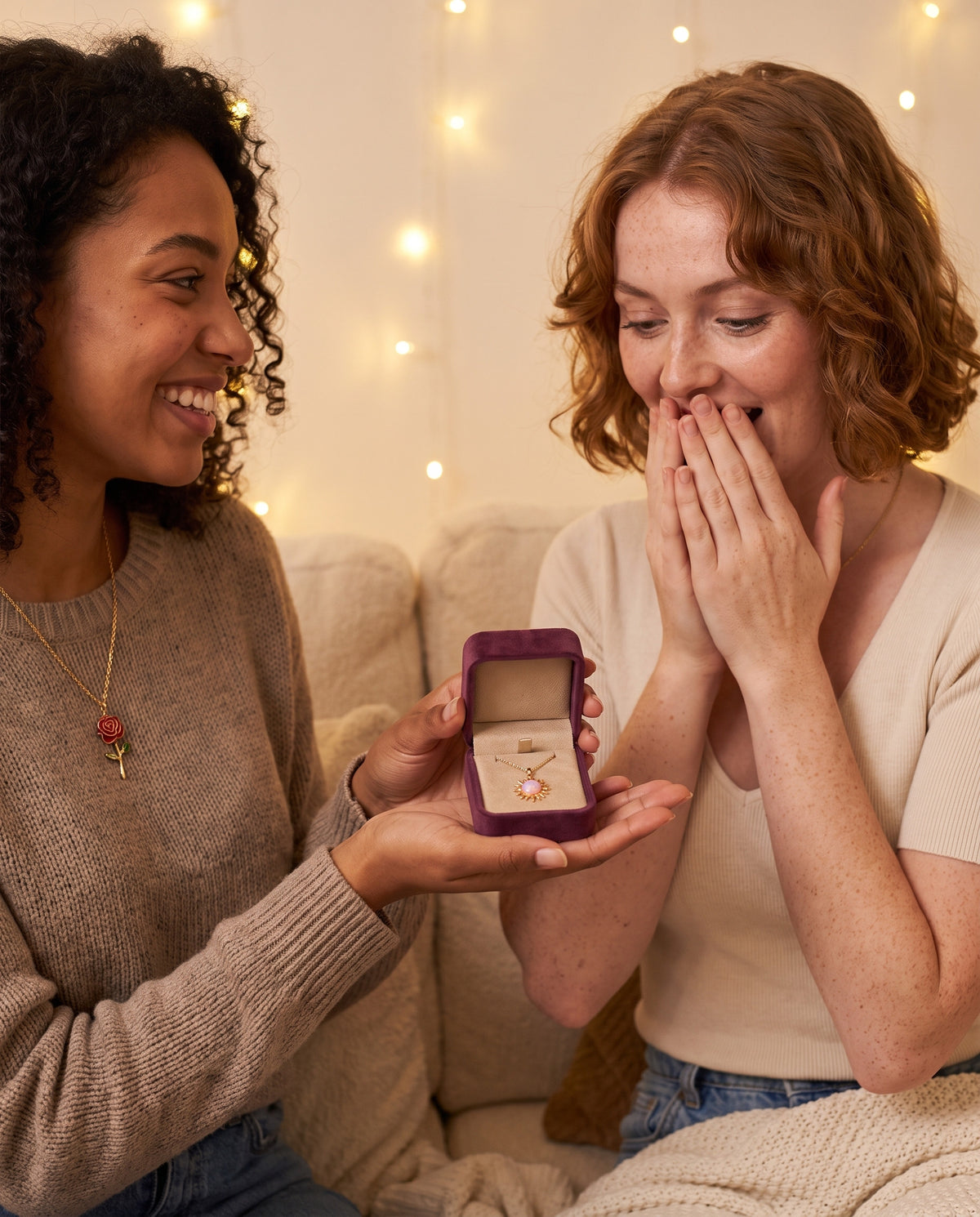 Two best friends on a sofa, one giving the other a Storyelle necklace in its velvet gift box
